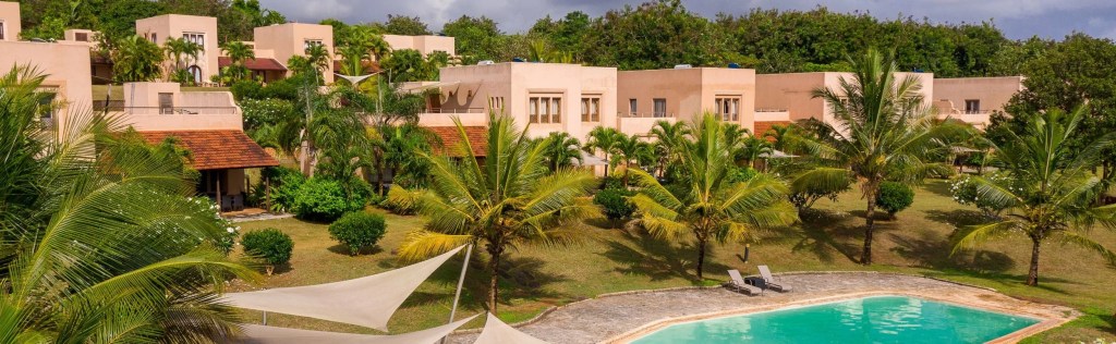 A scenic view of luxury villas surrounded by greenery at Vipingo Ridge, featuring a pool and palm trees under a partly cloudy sky.